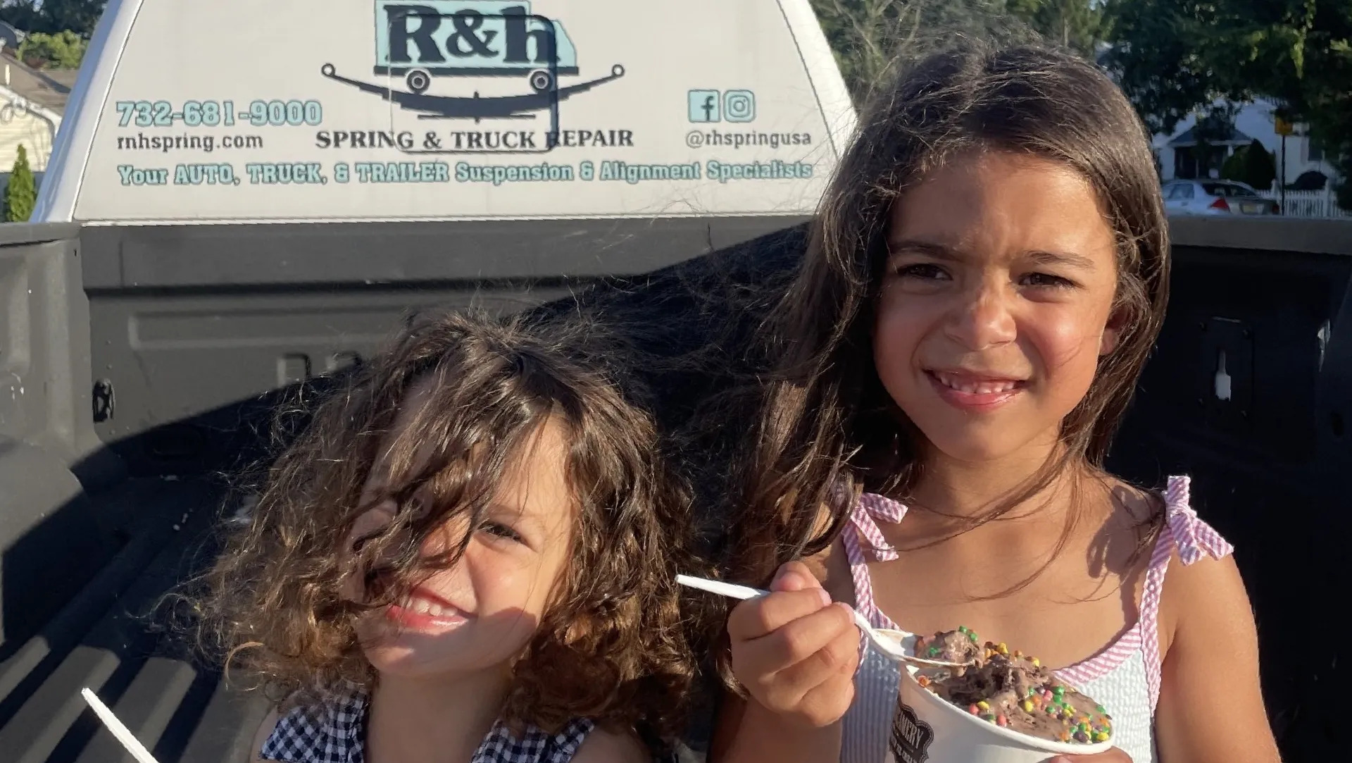 IMG_1141-1e76826e-a85923c9-1920w Two young girls sit in the bed of a truck eating ice cream. The truck tailgate has an R&H Spring & Truck Repair sign. One girl is smiling with messy hair, and the other is holding a cup of ice cream with sprinkles.