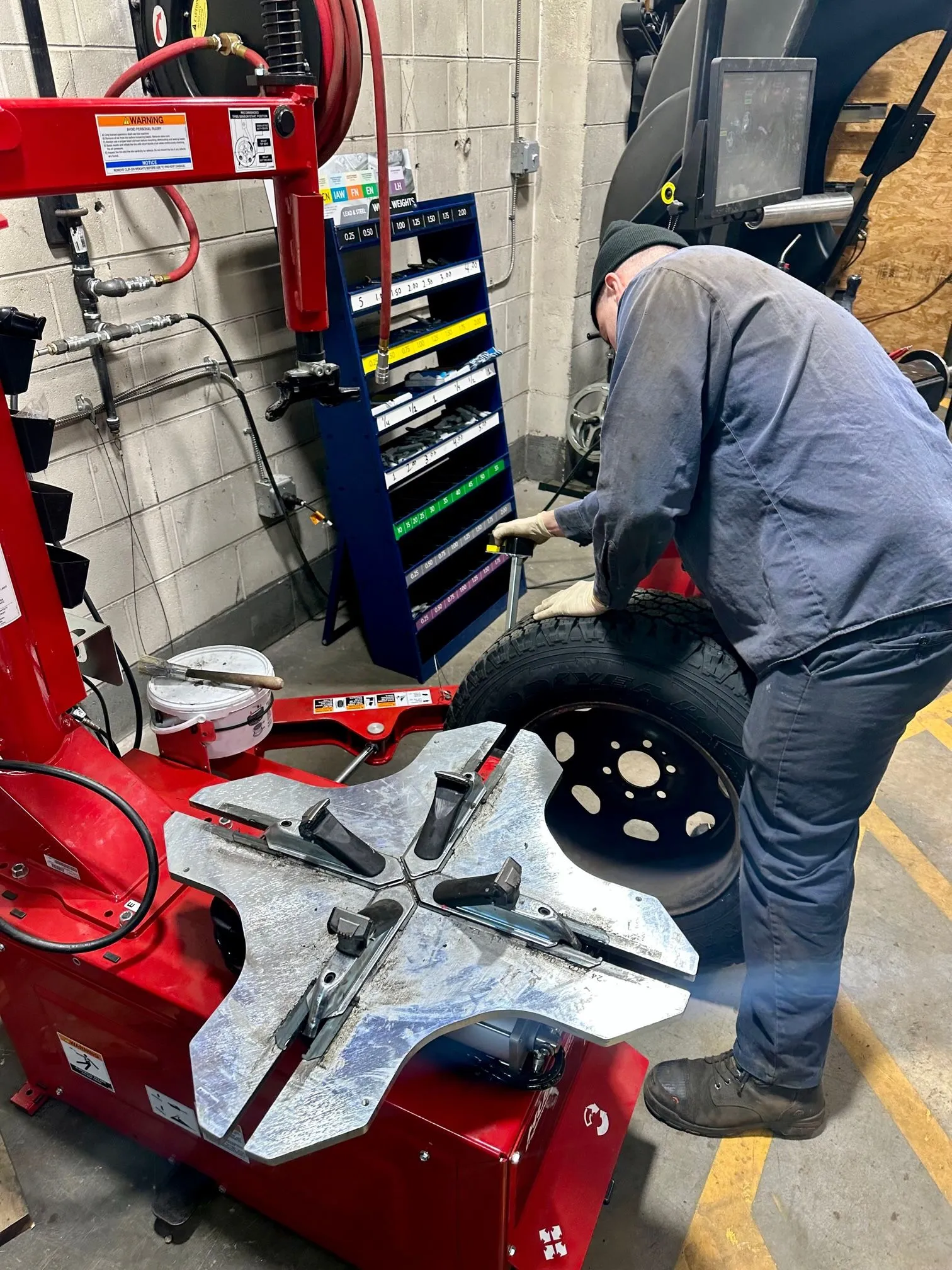 great-services A person in work clothes uses a tool to work on a car tire in an auto repair shop. Nearby are a red tire changing machine and an organized rack with colored storage bins.