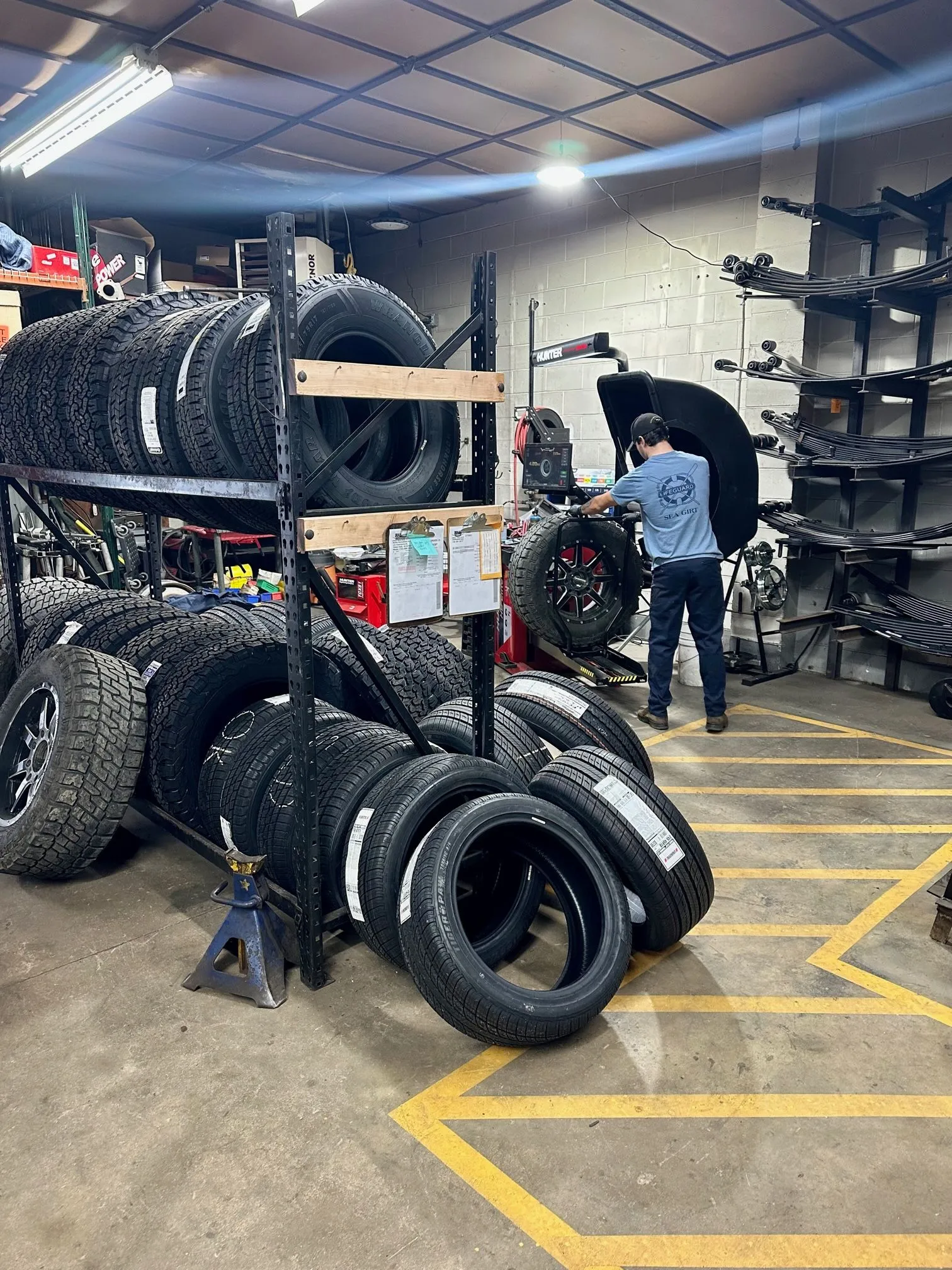 major-offered A person in a blue shirt works in a garage near a tire-changing machine, surrounded by stacked and standing tires on racks and the floor. The area is organized with yellow-striped lines on the ground.