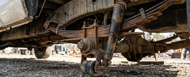 A photorealistic, high-end close-up of worn truck suspension components beneath a heavy-duty commercial pickup truck